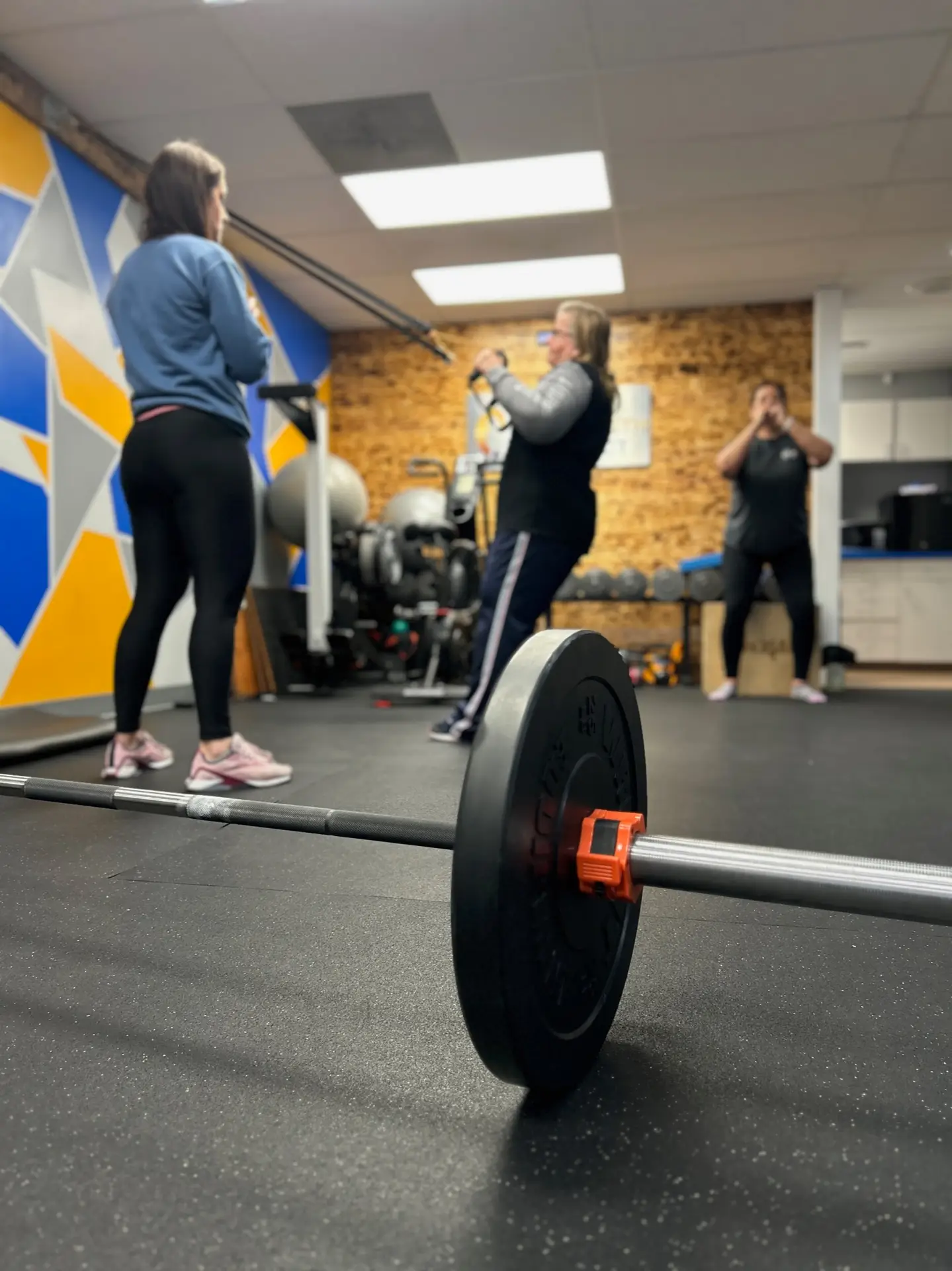 Barbell on the gym floor with three people in the background engaging in strength training exercises at Wellness Solutions Chiropractic in St. Louis County.